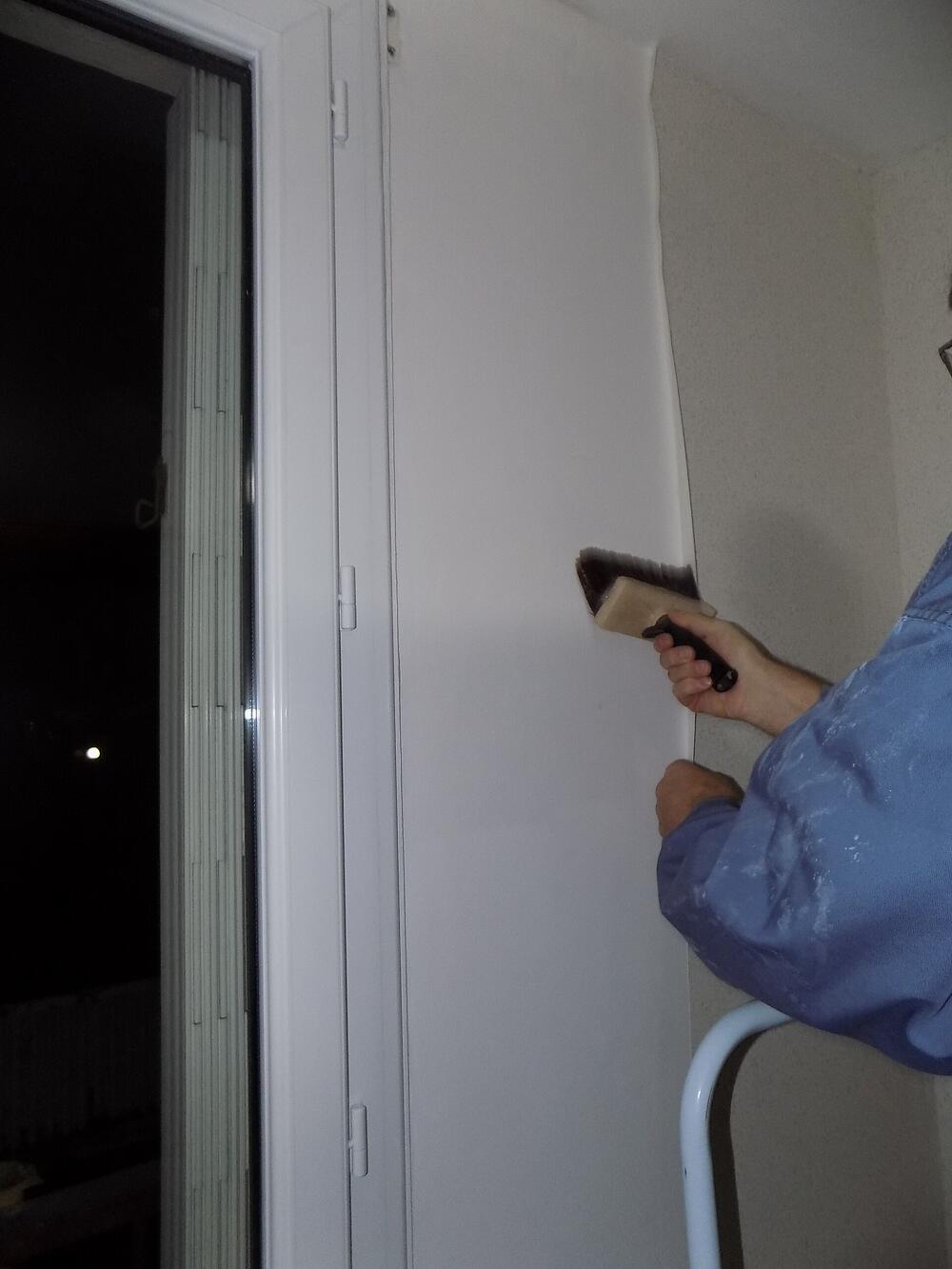 Een hand strijkt een baan behang glad met een behangborstel naast een raam